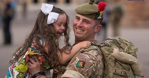 Returning soldier, still in uniform, hugs and lifts up their young daughter, both smiling. 