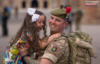 Returning soldier, still in uniform, hugs and lifts up their young daughter, both smiling.