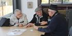 A male museum volunteer sits at a table with a couple, a man and a woman, discussing an item and documents that they have brought to the museum. The m
