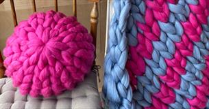 Pink chunky knitted cushion placed on a wooden chair, next to a blue and pink knitted blanket