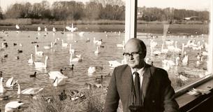 Sir Peter Scott sat in his Studio with a large window behind looking out onto the Rushy Lake which is filled with wild birds.