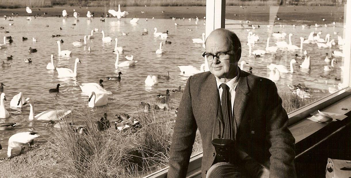 Sir Peter Scott sat in his Studio with a large window behind him looking out onto the Rushy Lake