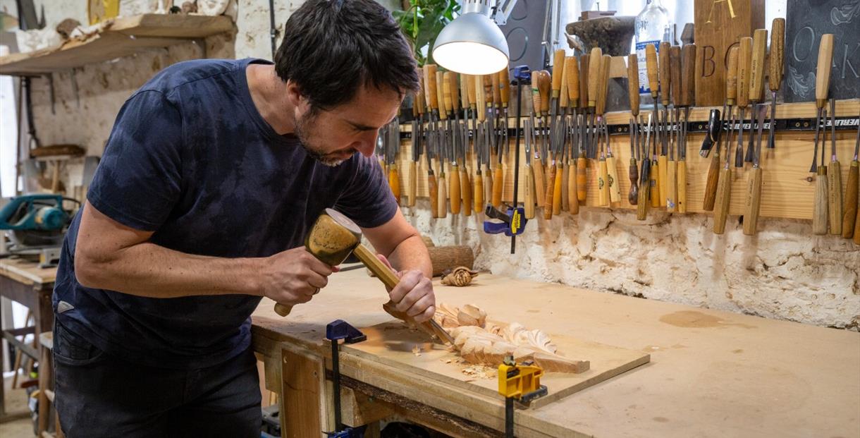 A person at a workbench using a hammer and chisel to carve a leaf design in wood