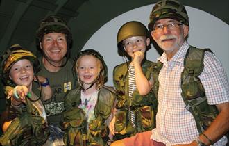 A family sitting inside one of the museum exhibits, a mock-up of a D-Day Horsa Glider. They are all wearing helmets and webbing from the museum's dres
