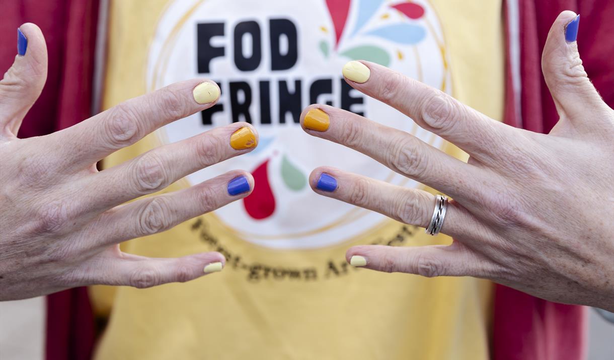 Image of hands with different coloured nail varish on finger nails. In the background is a t-shirt saying 'FOD FRINGE - a Forest Grown Festival'.