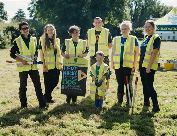 A group pic of 7 Attendees from last year's bogging event holding a sign saying I got mucky in the Bog.