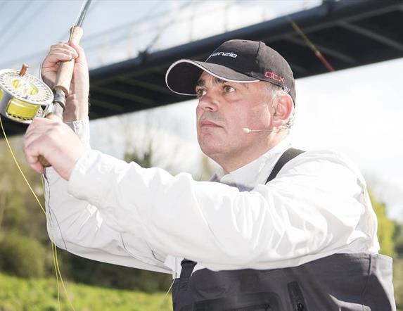 A man wearing a black cap and white shirt is fly fishing near a bridge. He holds a fishing rod with a reel of yellow fishing line, looking focused. A