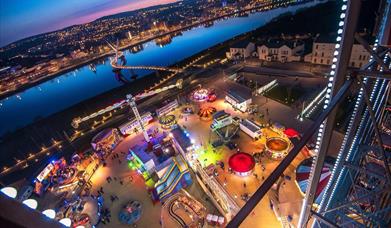 Birds eye view of a funfair beside a river at night-time.