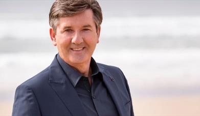 Daniel O'Donnell pictured in a black suit and shirt, smiling in front of a blurred background