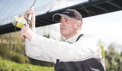 A man wearing a black cap and white shirt is fly fishing near a bridge. He holds a fishing rod with a reel of yellow fishing line, looking focused. A
