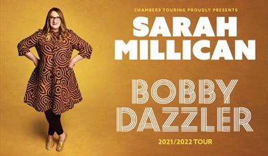 Sarah Millican standing with her hands on her hips in a loudly patterned dress, beside her in large text- 'SARAH MILLICAN- BOBBY DAZZLER' against a wa