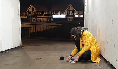 Man kneels on ground in a tunnel with houses in the distance. He appears to be operating some digital equipment.