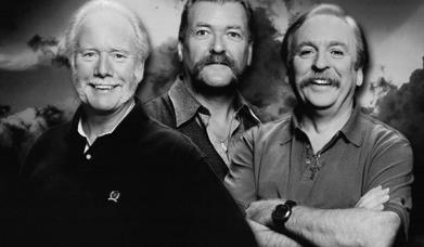 A black and white closeup  photo of The Wolfe Tones, smiling, with a dark, cloudy background