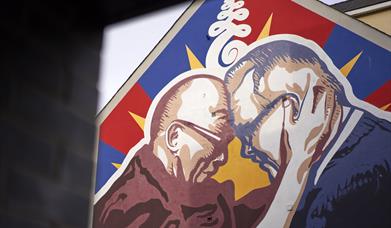 This is an image of a mural on a gable wall in Great James Street. The mural is of the Dalai Lama and Richard Moore. In the picture, their foreheads a