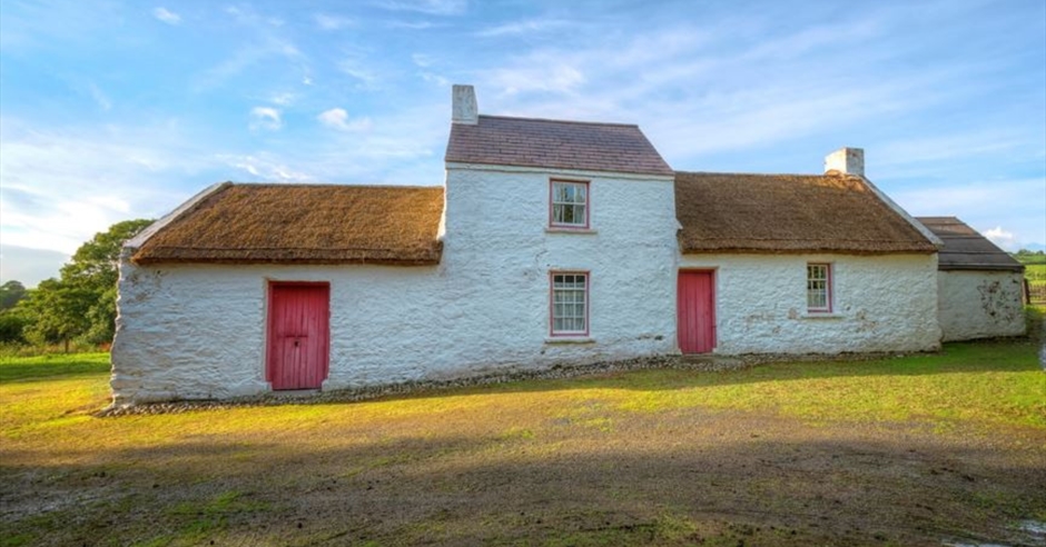 The historic Wilson Ancestral Home near Strabane, Northern Ireland, a white traditional cottage with a thatched roof, surrounded by green fields.