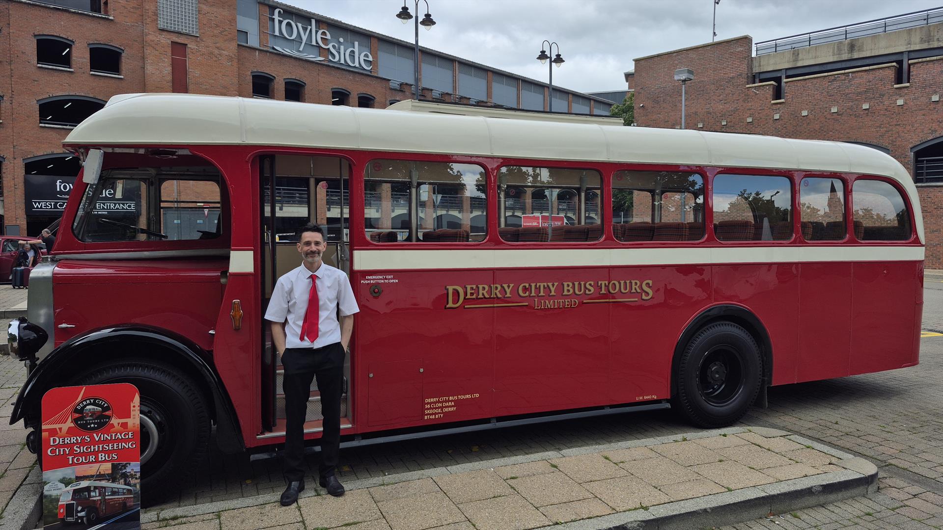 Vintage red and cream Derry City Bus Tours bus, driver/guide standing beside the entrance and a promotional sign at the front.