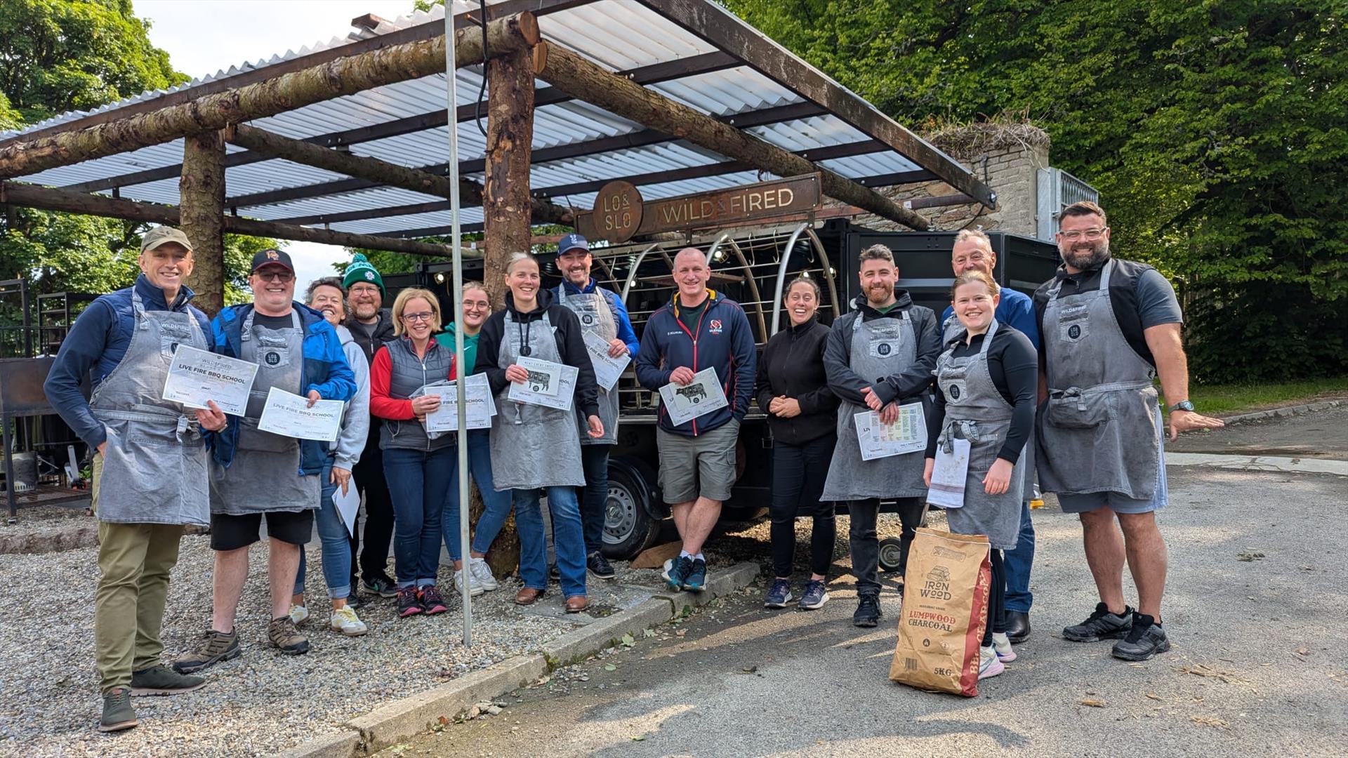 A group of fire school guests smiling in front of fire kitchen holding their certificates.
