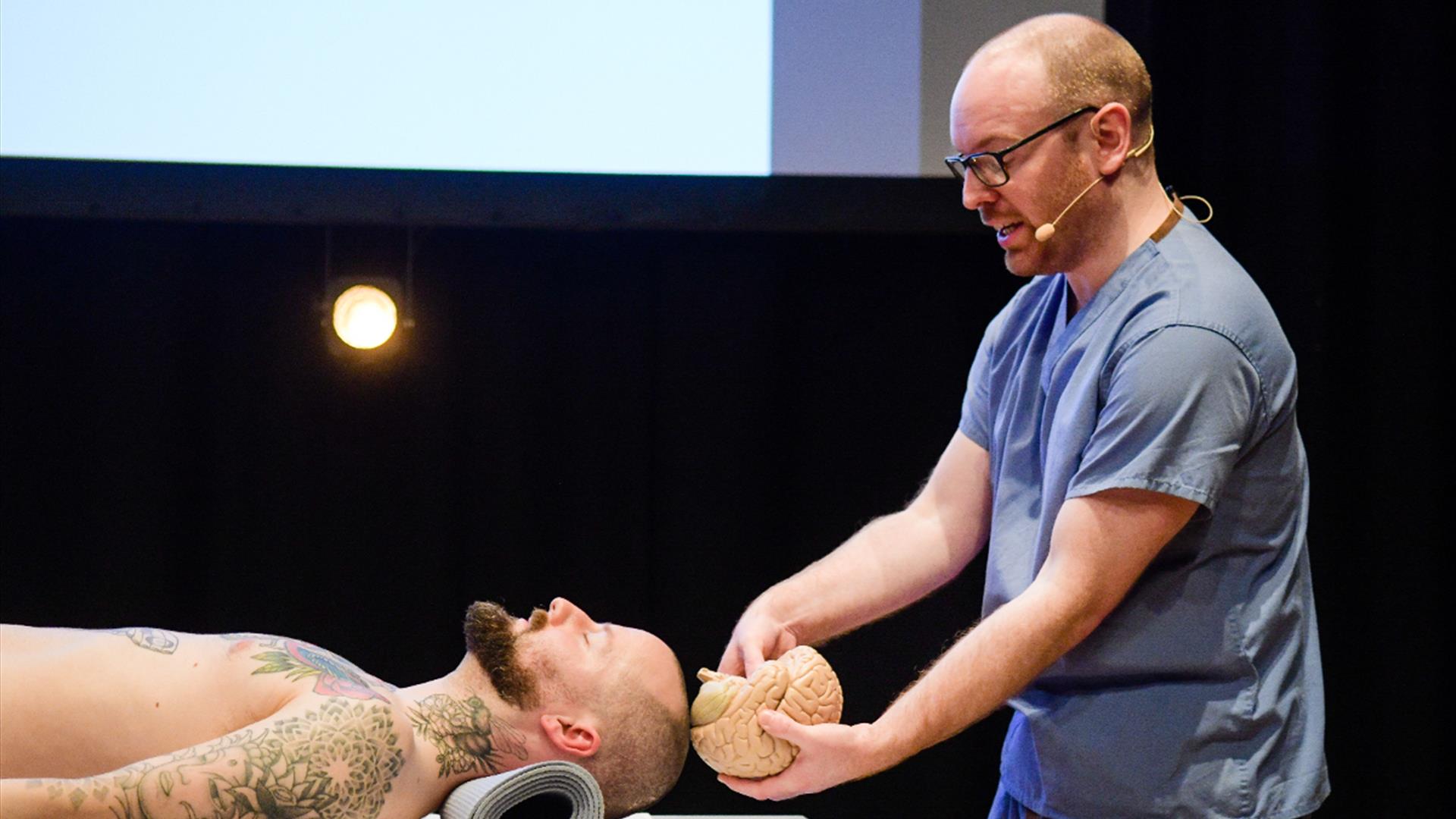 A male pathologist in blue scrubs with a beard and glasses holds a model brain  up to the skull of a male actor portraying a 'living' dead body.