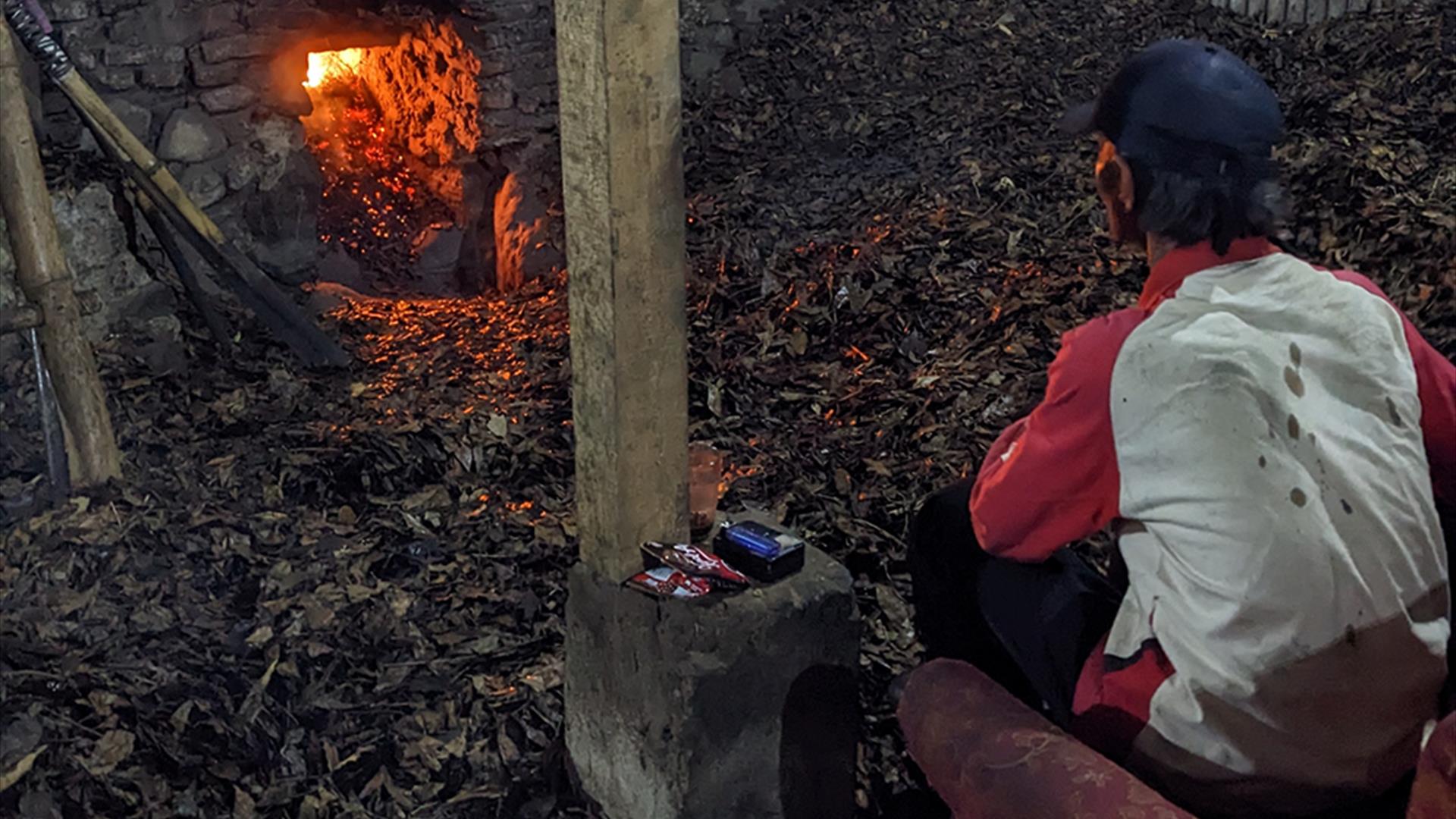 Image of a clove farmer watching a fire. He is sitting amongst leaves, and the fire can be seen reflecting from his face.