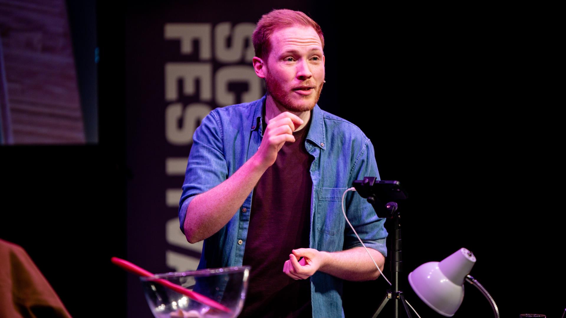 A man in a blue shirt and maroon t-shirt on stage with a mixing bowl Infront of him, clearly discussing a point.