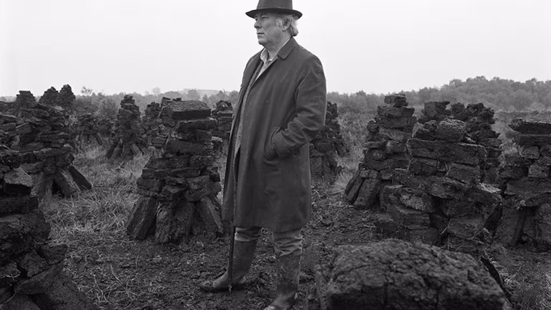 A black and white photograph of Irish poet Seamus Heaney in a hat and long coat, surrounded by stacks of peat in a field