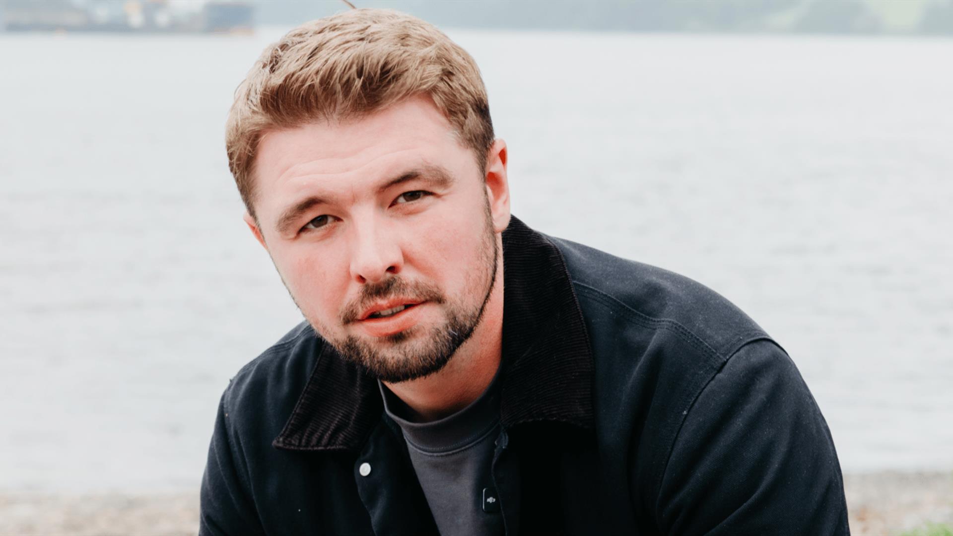 Jordan O'Keefe wearing a black jacket looking to the camera. He is on a beach by the sea.