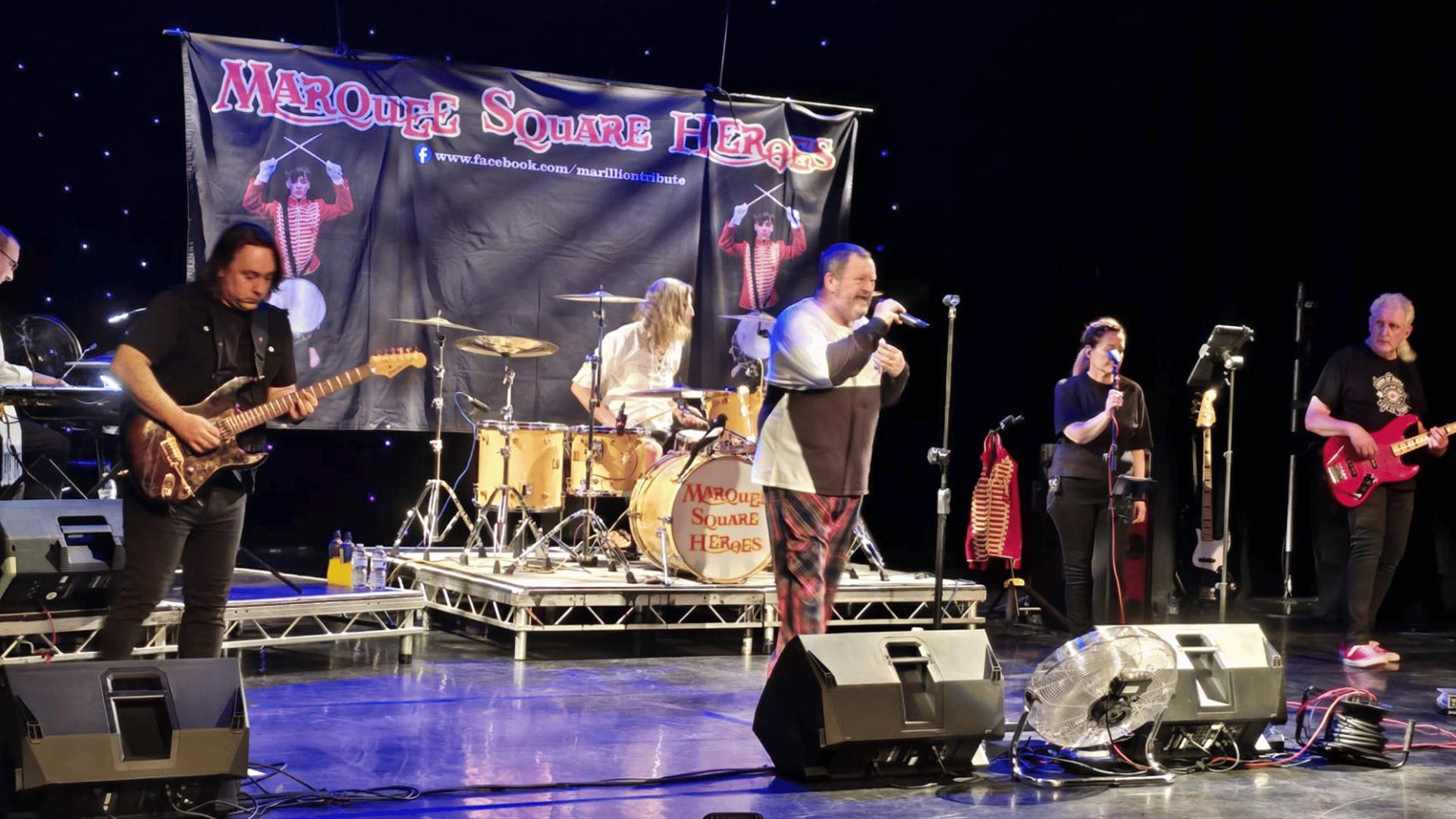 Marquee Square Heroes performing on a stage with guitars, drums, and microphones; a banner behind them reads "Marquee Square Heroes" and bright spotli