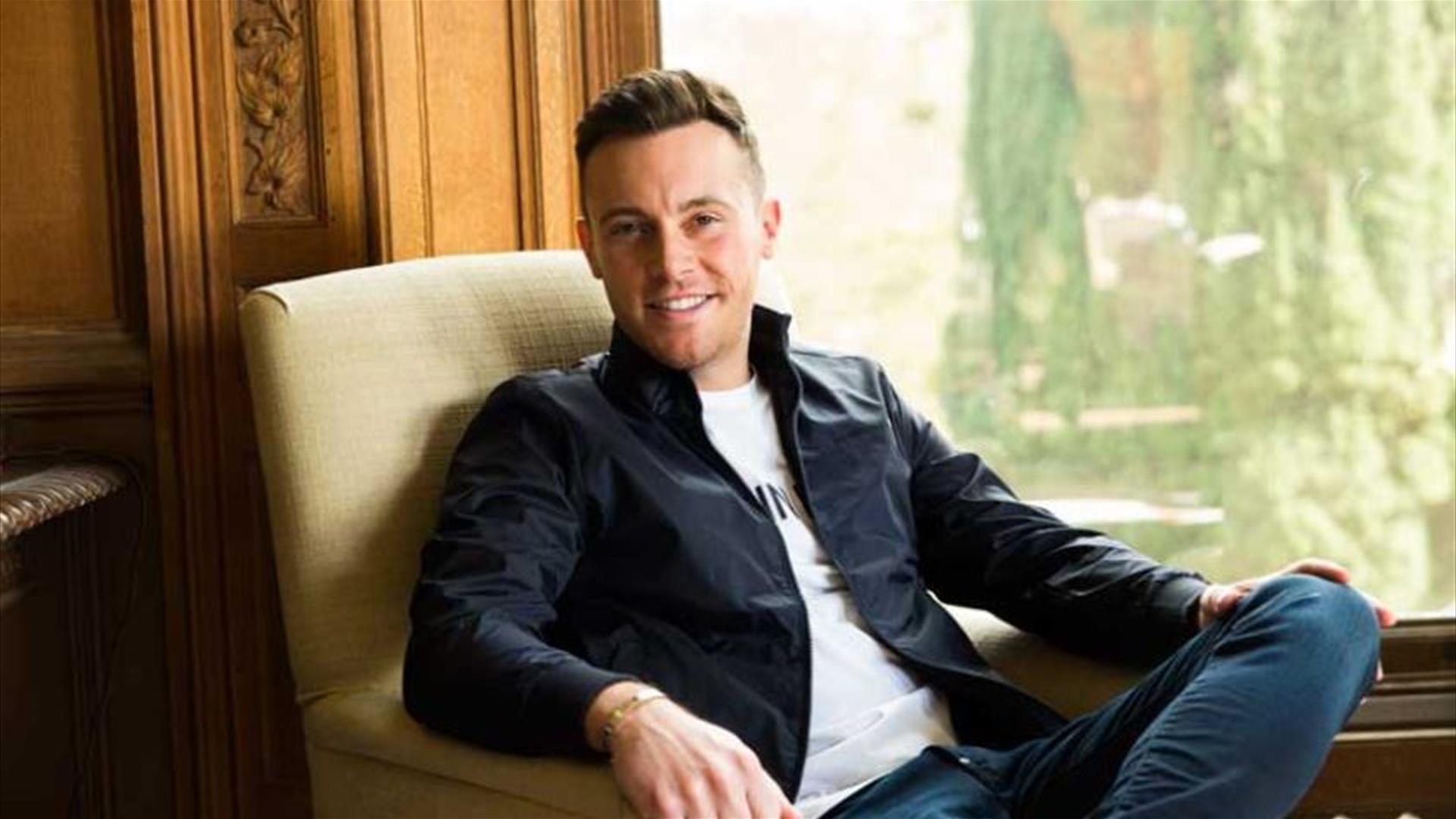 Nathan Carter, sitting and smiling leisurely in an armchair, in front of a window  with a view of greenery.