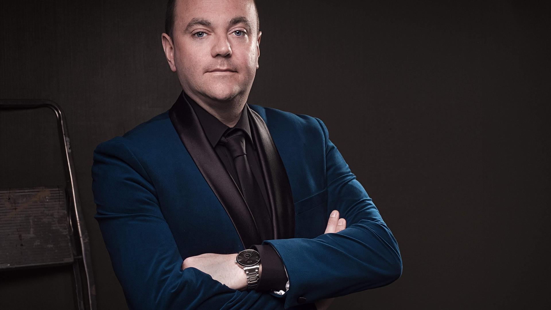 Portrait of Patrick Feeney, a confident man in a blue velvet blazer with black satin lapels, crossing his arms against a dark background. He wears a b