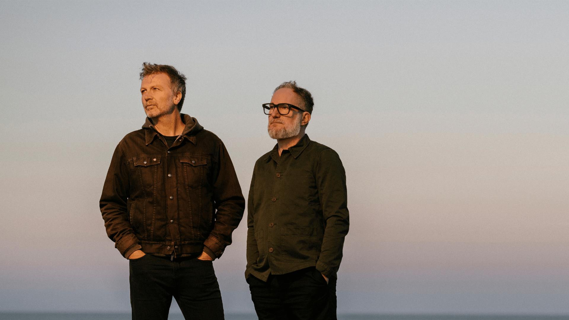 The members of Pilgrims stand on a grassy cliff near the sea, looking out at the sky at dusk.