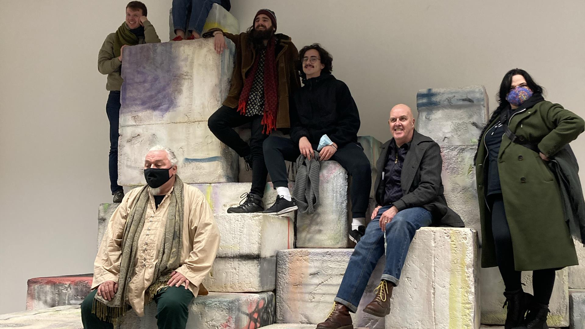 Man with long grey hair faces audience of people sitting on chairs. Man sits on what looks like cement blocks with graffiti covering them, inside a ro