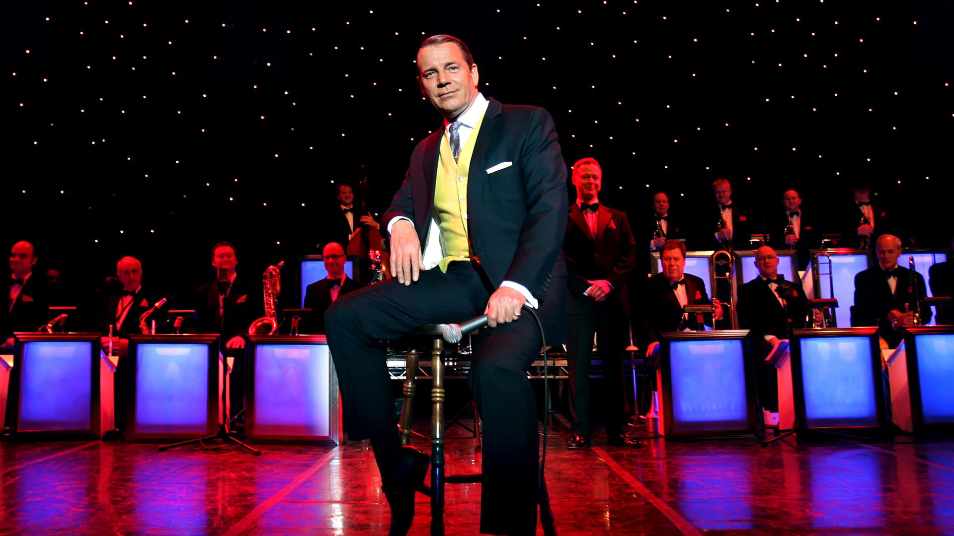 A stylish Stephen Triffitt as Frank Sinatra, seated centre stage in a classic suit and tie with a yellow waistcoat. The scene includes a live band in