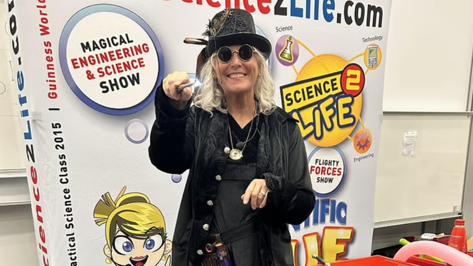 A person with curly blonde hair wearing glasses and steampunki inspired garments including a top hat waves a wand towards the camera.