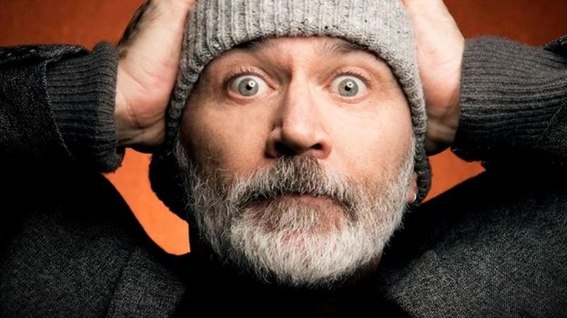 Tommy Tiernan wearing a grey hat, with his hands n his head- looking mildly exasperated against a burnt orange background.