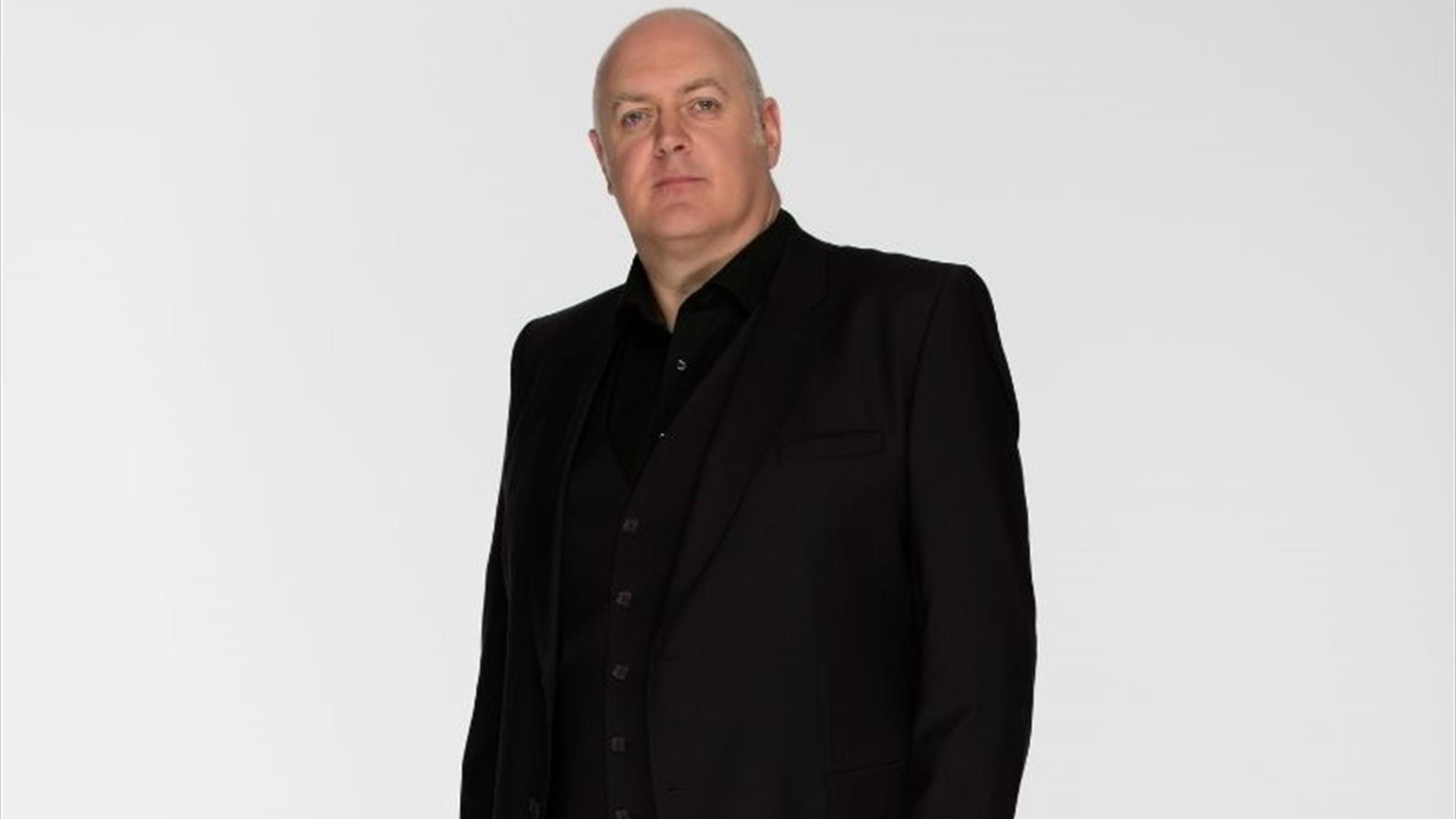 Dara O'Briain, sporting a neutral expression, standing in the centre in all black, against a white background