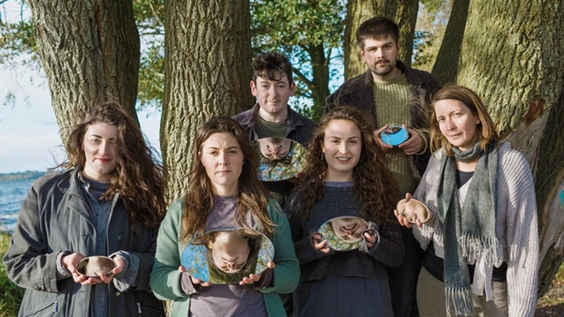 Hive Choir, a group of two young men and 4 young women, stand  by the shore with trees in the background, each  holding small oval mirrors