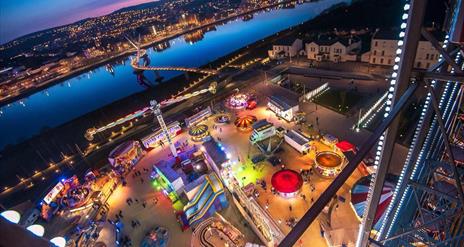 Birds eye view of a funfair beside a river at night-time.