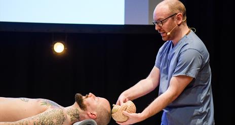 A male pathologist in blue scrubs with a beard and glasses holds a model brain  up to the skull of a male actor portraying a 'living' dead body.