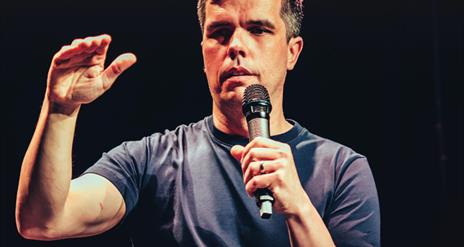 Chris kent holds a mic as he performs his arm slightly bent with his hand raised