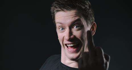 Daniel Sloss, grinning in an animated fashion, with the silhouette of his middle finger in front of his face. Against an all-black background.