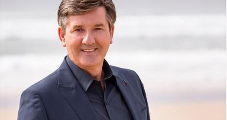 Daniel O'Donnell pictured in a black suit and shirt, smiling in front of a blurred background