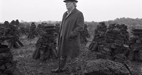 A black and white photograph of Irish poet Seamus Heaney in a hat and long coat, surrounded by stacks of peat in a field