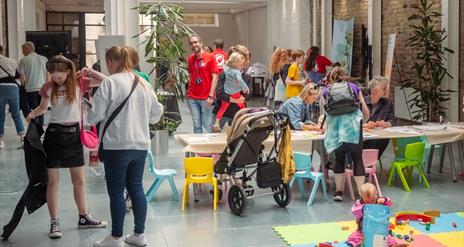 A busy indoor scene  of children and families drawing at a table in a gallery
