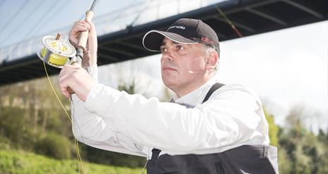 A man wearing a black cap and white shirt is fly fishing near a bridge. He holds a fishing rod with a reel of yellow fishing line, looking focused. A