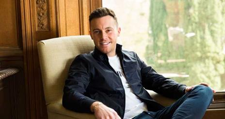 Nathan Carter, sitting and smiling leisurely in an armchair, in front of a window  with a view of greenery.