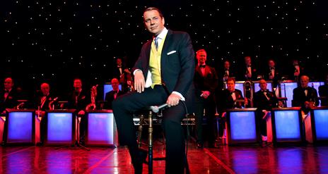 A stylish Stephen Triffitt as Frank Sinatra, seated centre stage in a classic suit and tie with a yellow waistcoat. The scene includes a live band in