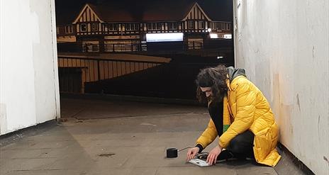 A young man with long hair in a yellow coat crouches in an underpass and operates  a keyboard attached  to a speaker.