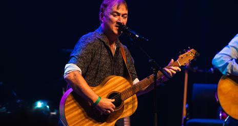 Image shows Brian standing, holding a guitar