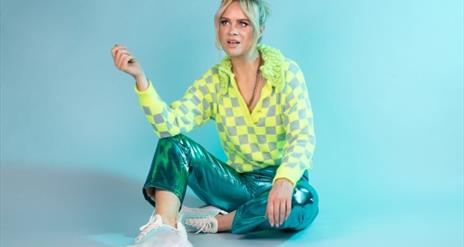 Joanne sitting casually on the floor in a bright yellow and turquoise outfit, against a cool green/blue background.