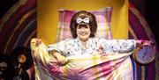 A cheerful character with voluminous hair and a bow smiles brightly from a yellow bed with colourful plaid bedding, holding an alarm clock and surroun